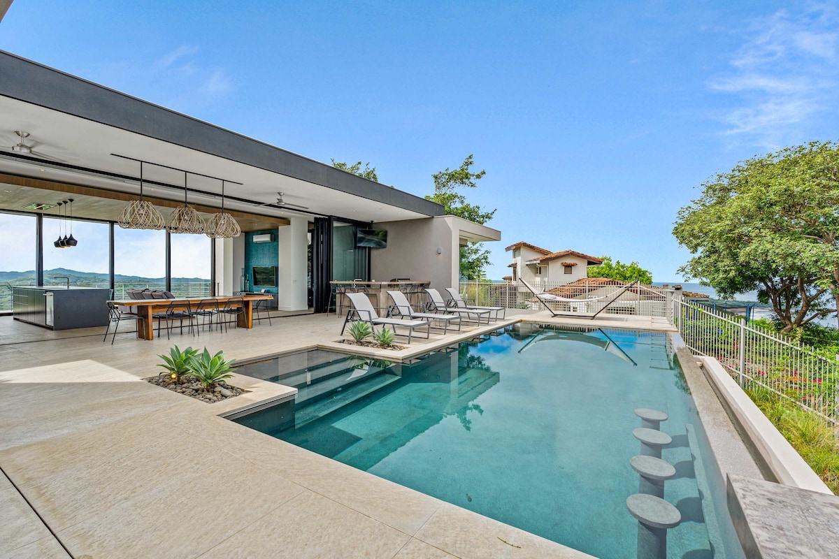 A modern outdoor space features an infinity pool surrounded by comfortable loungers. Nearby, a spacious dining table is visible, complemented by an open layout that connects to a shaded area with expansive glass doors. Lush greenery and coastal views enhance the overall ambiance.