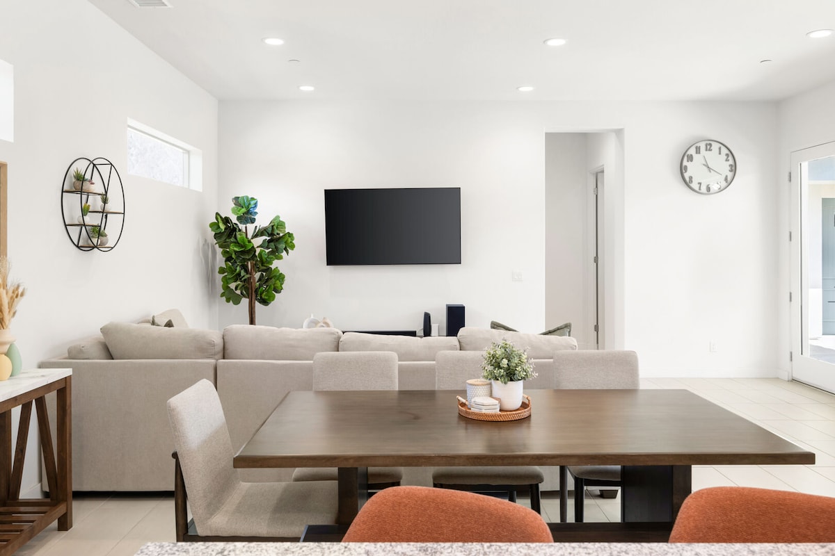 An open concept living area displays a comfortable sofa facing a wall-mounted television. A modern dining table is centrally located, surrounded by chairs. Natural light fills the space through large windows, complemented by indoor plants and neutral-colored decor.
