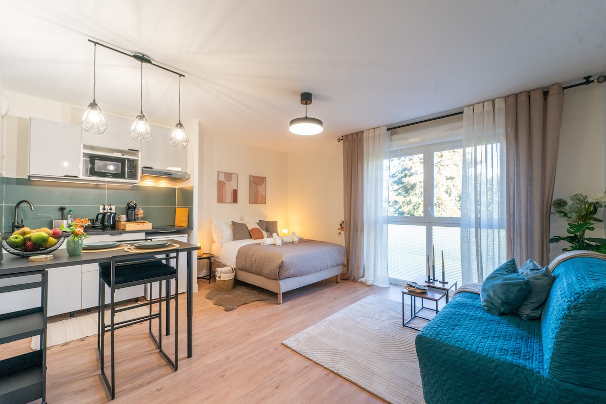 A modern living area combines a well-equipped kitchen with a dining area, and a cozy seating arrangement. A spacious bed is visible alongside the window, allowing natural light to filter through sheer curtains. Decorative plants enhance the inviting ambience of the room.
