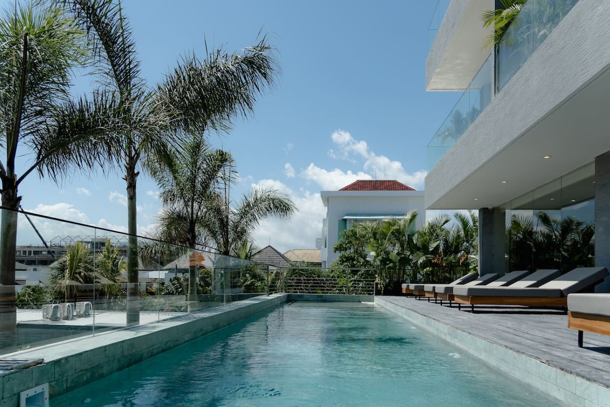 A serene pool area is framed by lush palm trees, providing a tranquil escape. Sun loungers line the deck, inviting relaxation under the clear blue sky. The elegant building structure is seen in the background, showcasing modern architectural elements and large windows.
