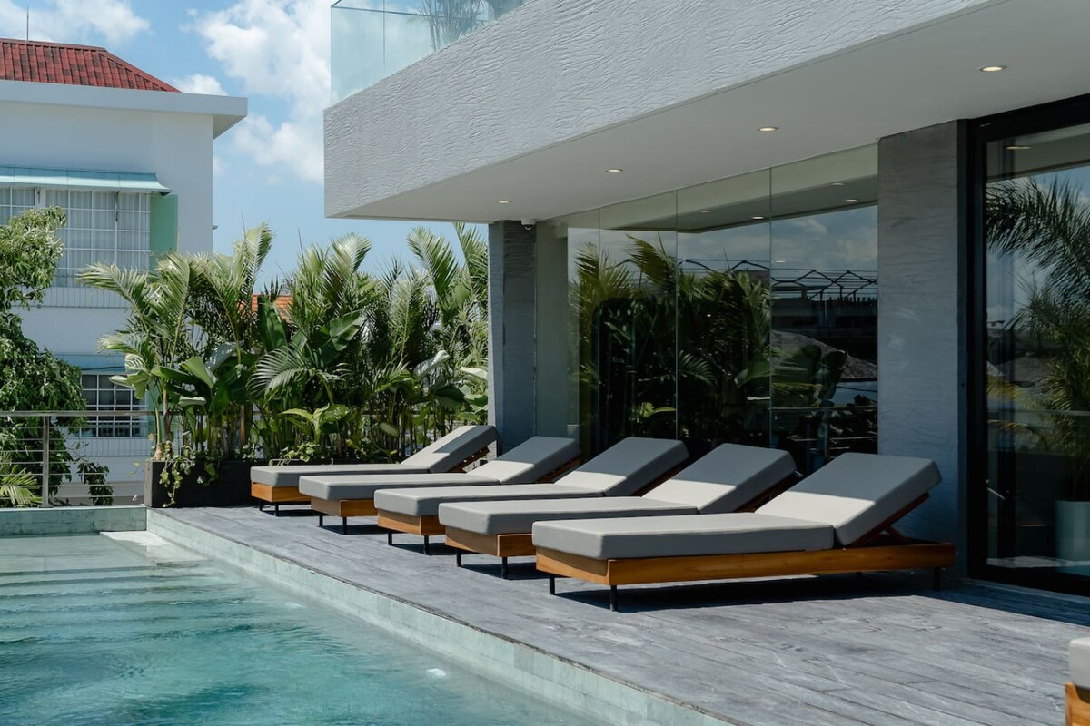 A sunlit outdoor area features several chaise lounge chairs arranged along a wooden deck beside a serene swimming pool. Lush greenery surrounds the space, creating a peaceful retreat ideal for relaxation.
