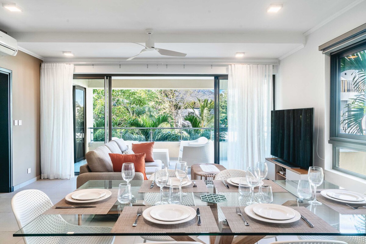 A dining area features a glass table set for six, with elegant dishware and glassware arranged neatly. Directly accessible through large glass doors, the living space reveals a cozy seating arrangement with soft furnishings and lush greenery visible outside.