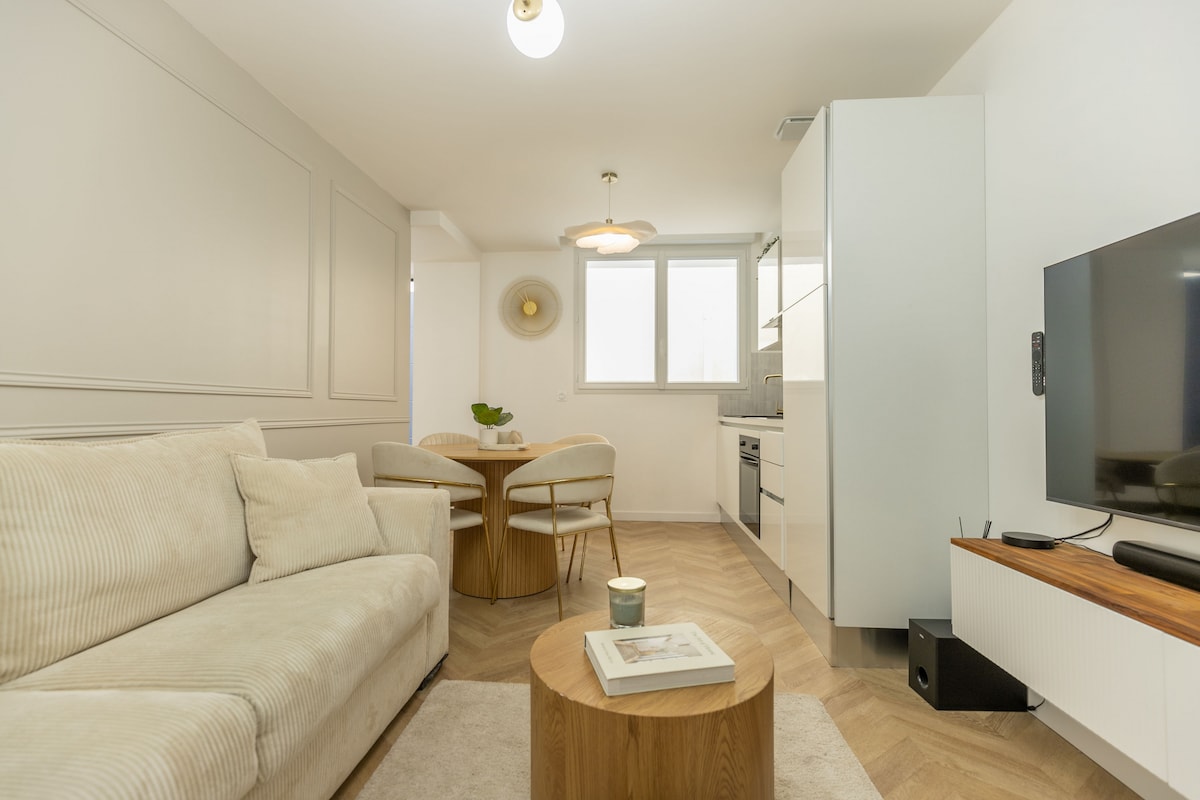 A light-filled living area features a comfortable beige sofa complemented by a round wooden coffee table. A small dining set with two chairs sits nearby, and a modern kitchen is visible in the background. A large television is mounted on the wall for entertainment.