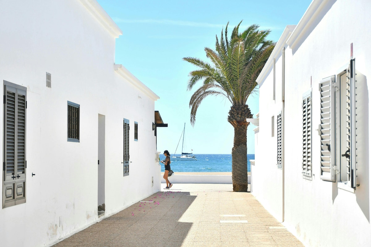 A narrow pathway is flanked by white buildings featuring wooden shutters. A palm tree stands prominently, silhouetted against the blue sea. In the background, a sailboat can be seen gently bobbing on the water, contributing to the coastal atmosphere.