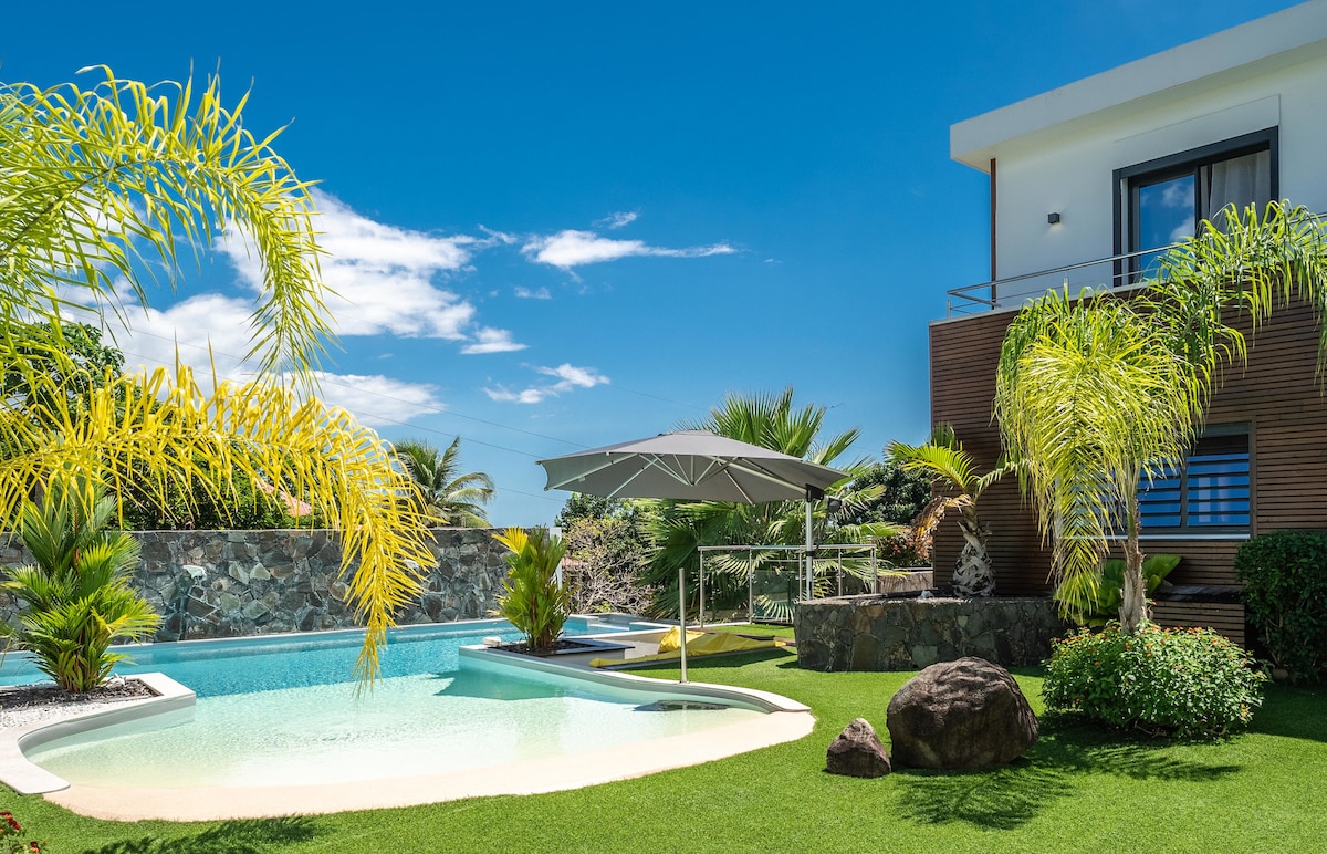 A private heated pool is surrounded by lush greenery and palm trees. An umbrella provides shade over a seating area next to the clear water, while a well-manicured lawn adds to the tranquil setting. The villa's modern exterior features large windows that enhance the outdoor view.