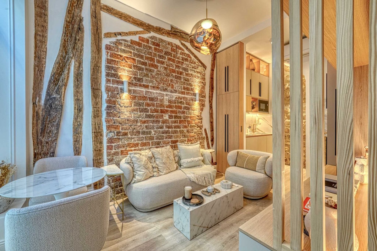 A cozy living area features a mix of modern and rustic elements, highlighted by a brick wall and stylish furnishings. A marble coffee table sits centrally, accompanied by plush sofas. Soft lighting enhances the inviting ambiance, while a glimpse of the kitchen is visible in the background.