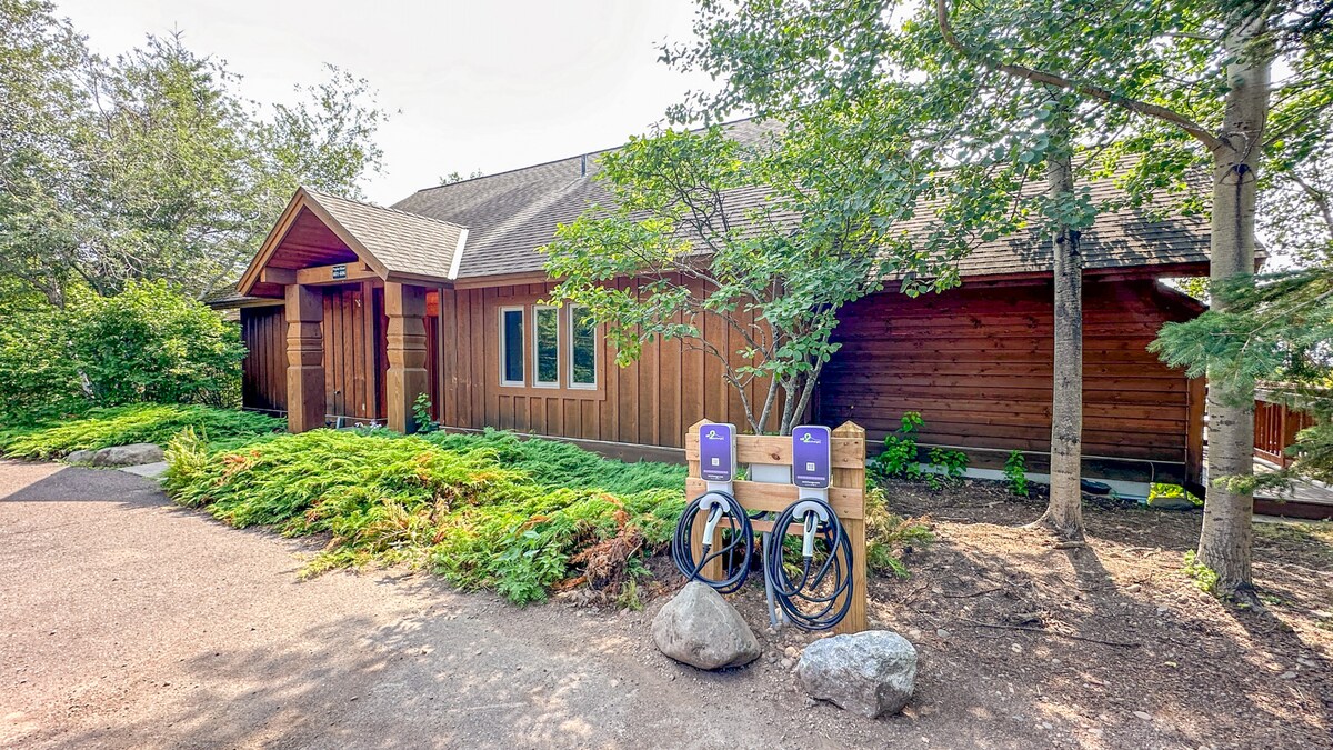 The exterior of the condo is framed by trees, showcasing a wooden facade and a welcoming entrance. Electric vehicle charging stations are positioned near the pathway, alongside natural landscaping with rocks and greenery that enhances the setting.