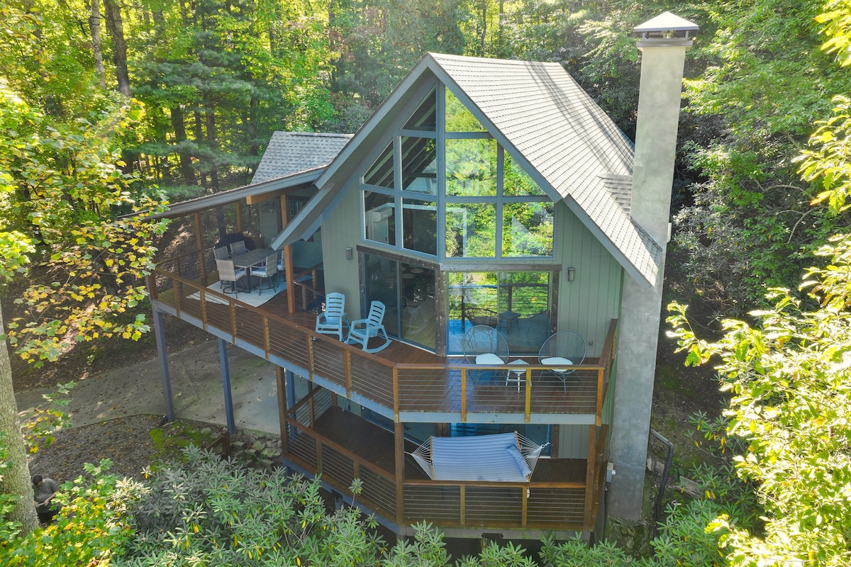 The retreat is positioned amidst trees with an elevated deck featuring outdoor seating and a hammock. The structure showcases a large wall of windows, allowing natural light to illuminate the interior, while a stone chimney enhances its rustic charm.