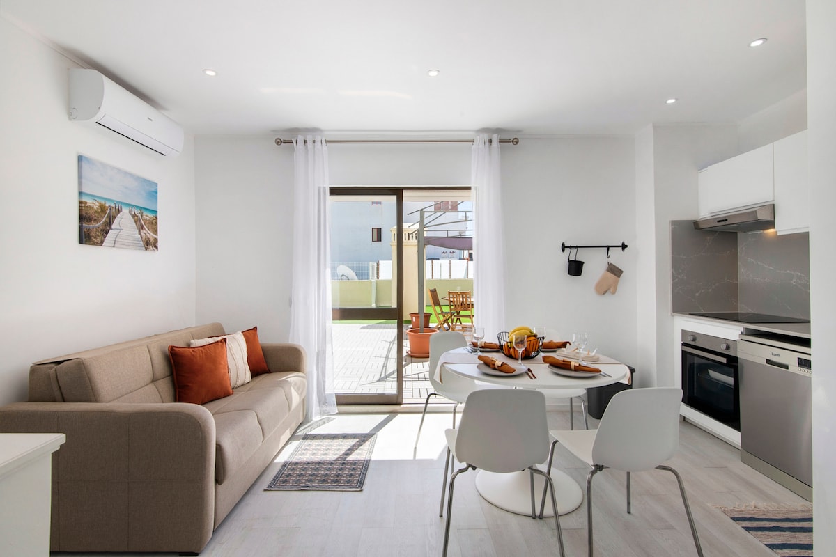 A bright living area is presented with a comfortable sofa and a dining table set for meals. Natural light fills the space through sliding glass doors, which lead to an outdoor area. Air conditioning is also visible, enhancing comfort during warm days.