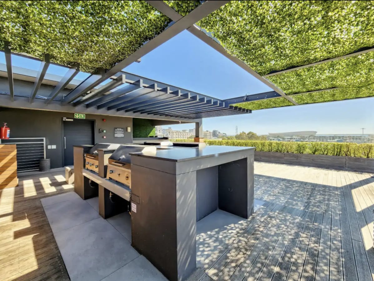 An outdoor grilling area is presented with built-in barbecues beneath a canopy of greenery. The spacious wooden deck allows for ample movement, with views of the surrounding landscape visible in the distance. Natural light floods the area, creating an inviting space for gatherings.