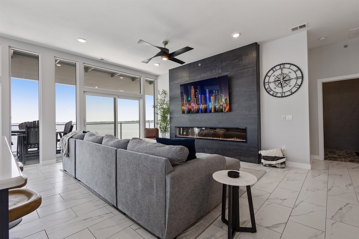 A modern living area features a spacious gray sectional couch facing a sleek electric fireplace and a large wall-mounted television. Floor-to-ceiling windows provide extensive lake views and natural light, while a contemporary clock adds a stylish touch to the decor.