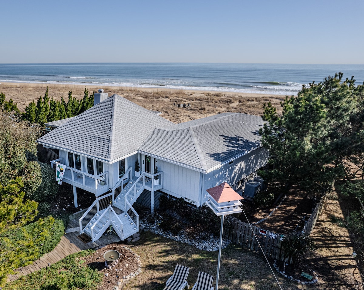 The exterior of the house features a multi-gabled roof and a spacious deck, surrounded by a sandy area. A few beach chairs are positioned nearby. The ocean extends in the background, with gentle waves lapping against the shoreline and clear blue skies overhead.