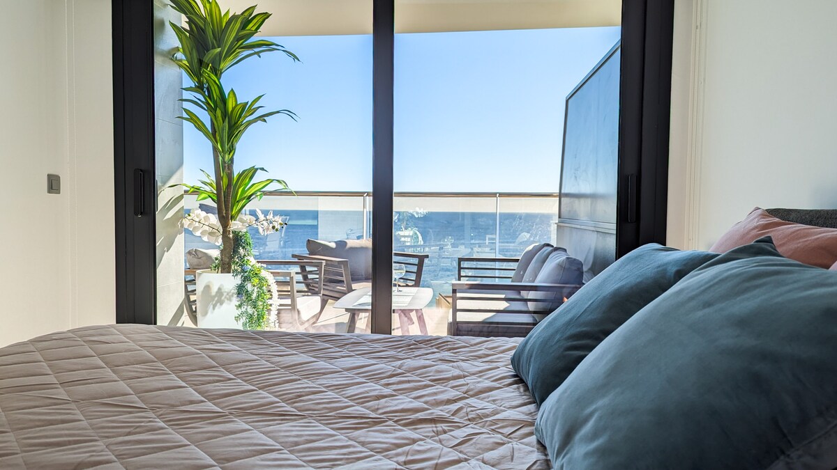 A spacious sleeping area features a neatly made bed with a quilted cover and accent pillows. Large glass doors offer stunning views of the sea and lead to a terrace, where outdoor furniture invites relaxation.