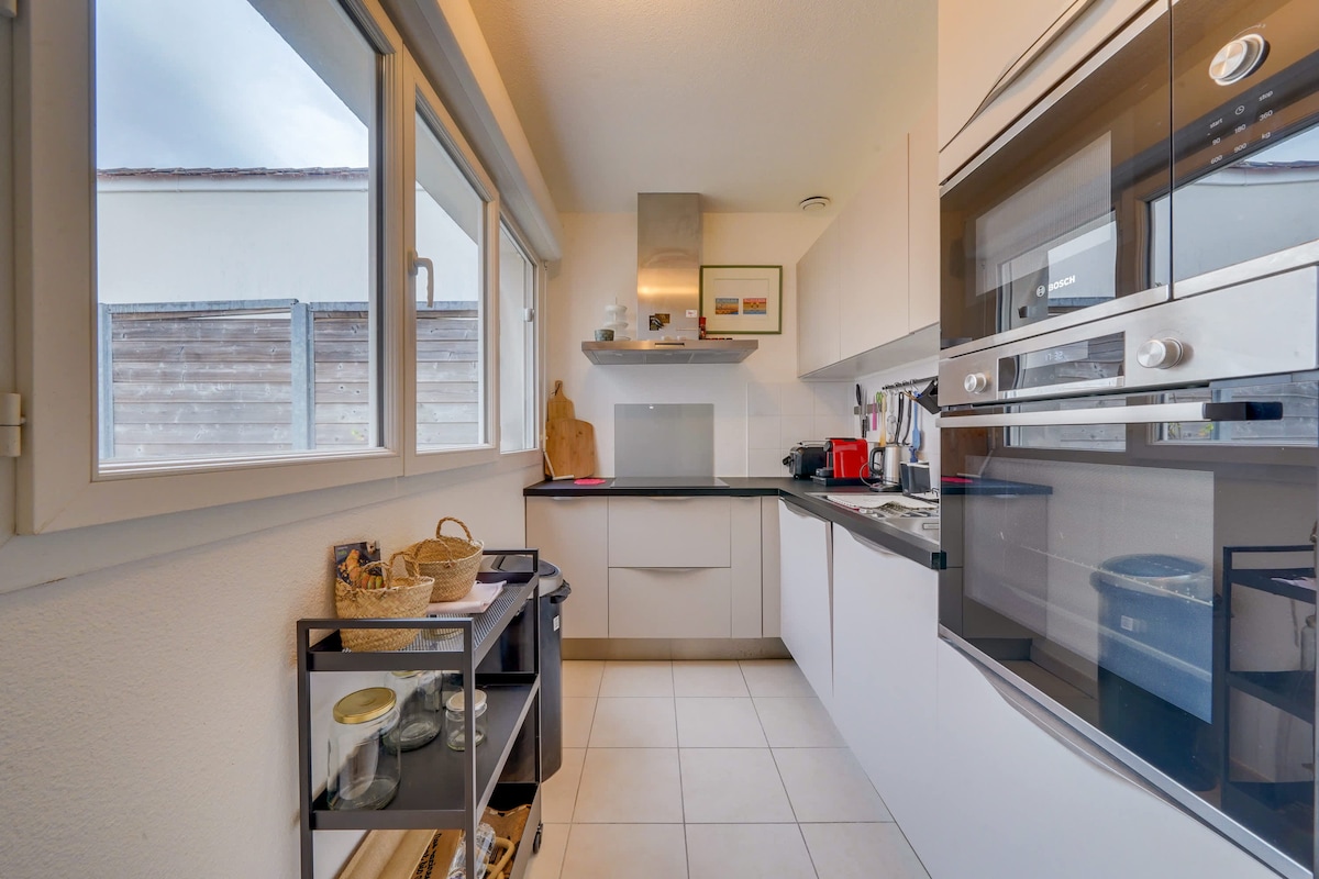 The kitchen features modern appliances including an oven, microwave, and a brightly lit countertop. Natural light enters through large windows, illuminating the space. A storage unit holds various kitchen items, with a sleek design complementing the overall decor.