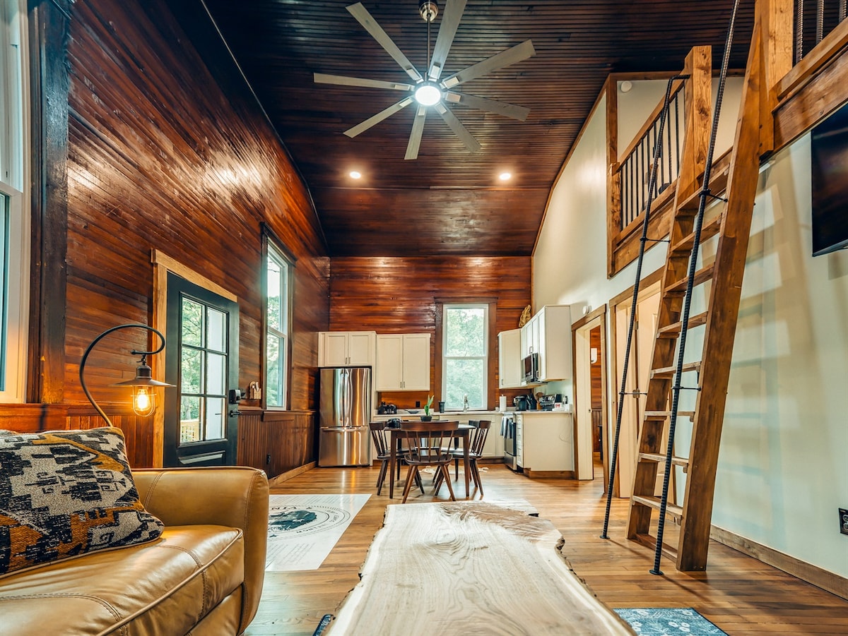 An open living area showcases rich wooden walls and ceiling, complemented by a modern kitchen in white cabinetry. A cozy seating area with a soft couch is positioned near a dining table. A wooden ladder leads to an upper loft, enhancing the spacious feel.