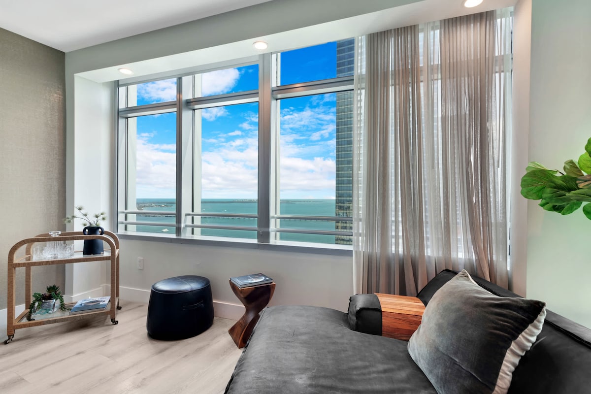 A modern seating area features large floor-to-ceiling windows offering views of the water and sky. Soft curtains frame the windows, allowing for natural light. A sleek sofa rests beside a small side table, complemented by a stylish bar cart with decorative items.