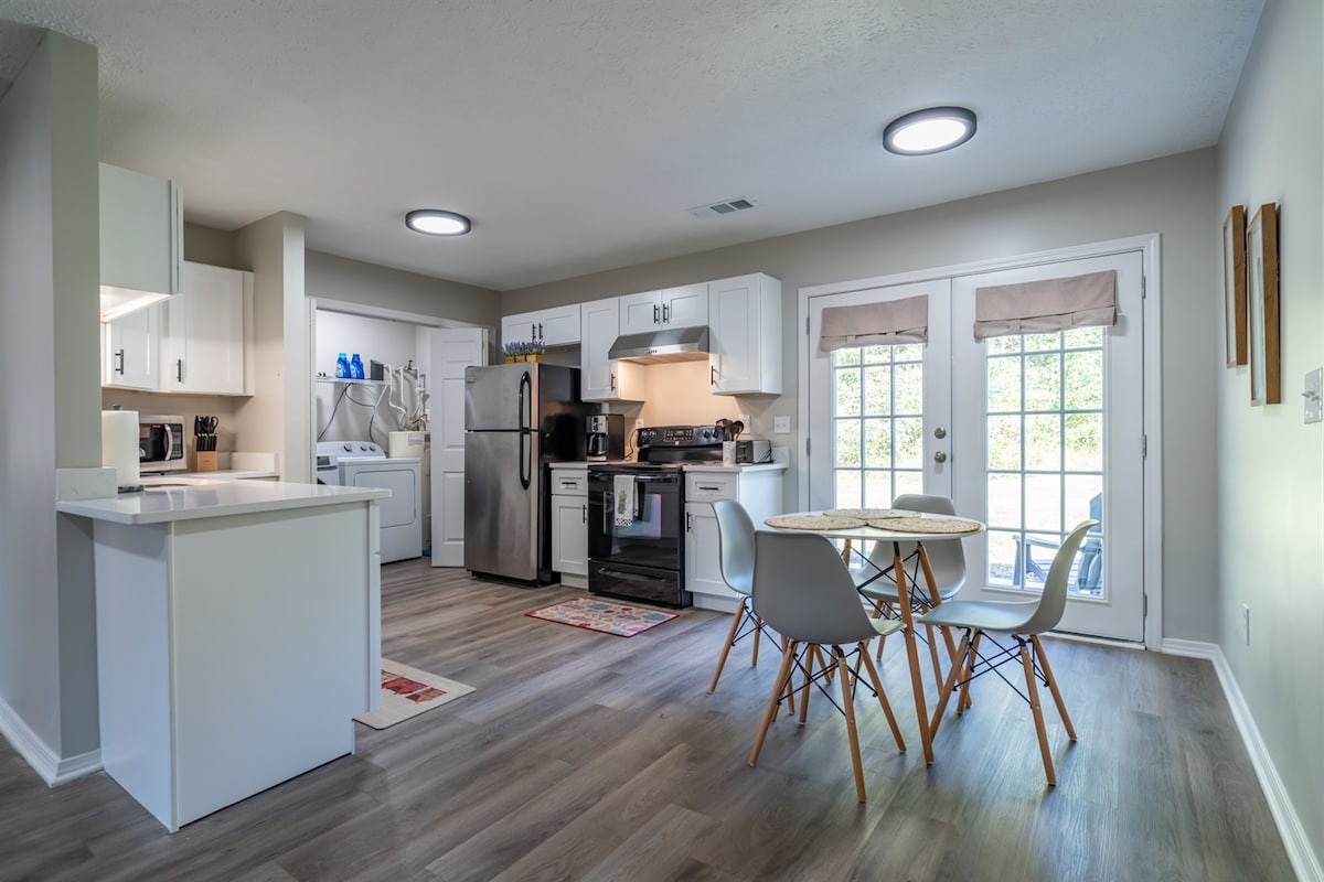 A modern kitchen and dining area is presented, featuring white cabinetry and stainless steel appliances. A dining table with four chairs stands near glass doors that provide natural light and access to the outside. Light-colored flooring complements the bright interior design.