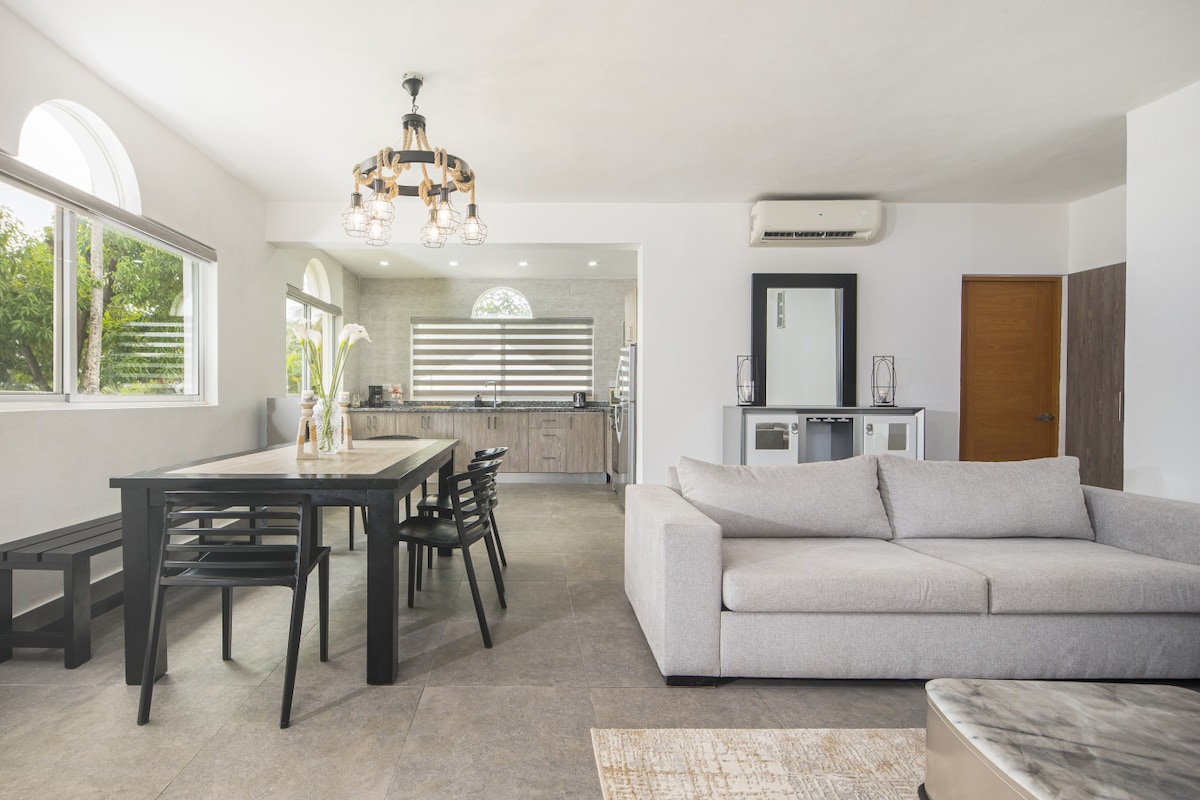 An open living area features a dining table with six black chairs and a spacious light grey sofa. Natural light fills the room through large arched windows, illuminating the modern kitchen area in the background. A sleek air conditioning unit is mounted on the wall.
