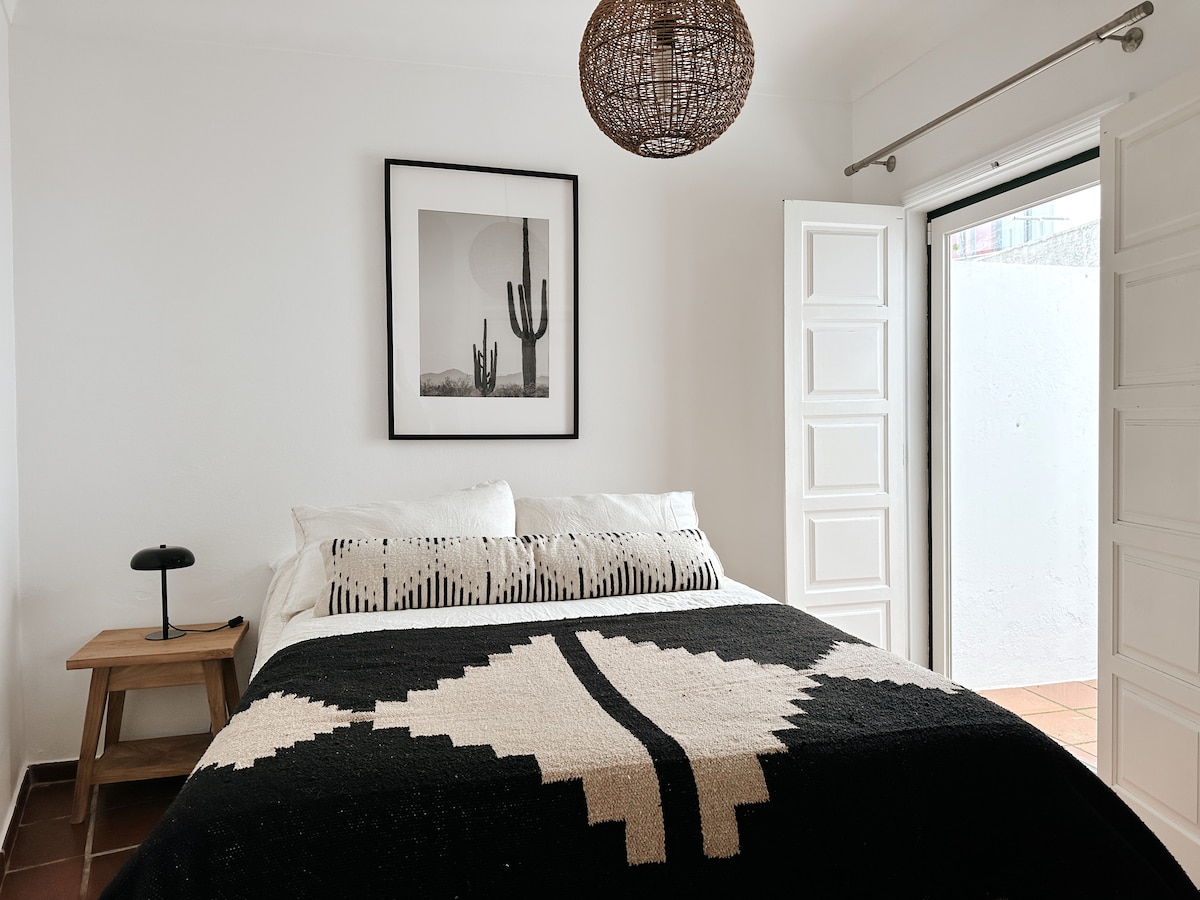 A cozy bedroom features a spacious bed adorned with a patterned black and white blanket. Natural light enters through open French doors, which lead to a small outdoor area. A minimalist bedside table with a lamp complements the room's contemporary design.