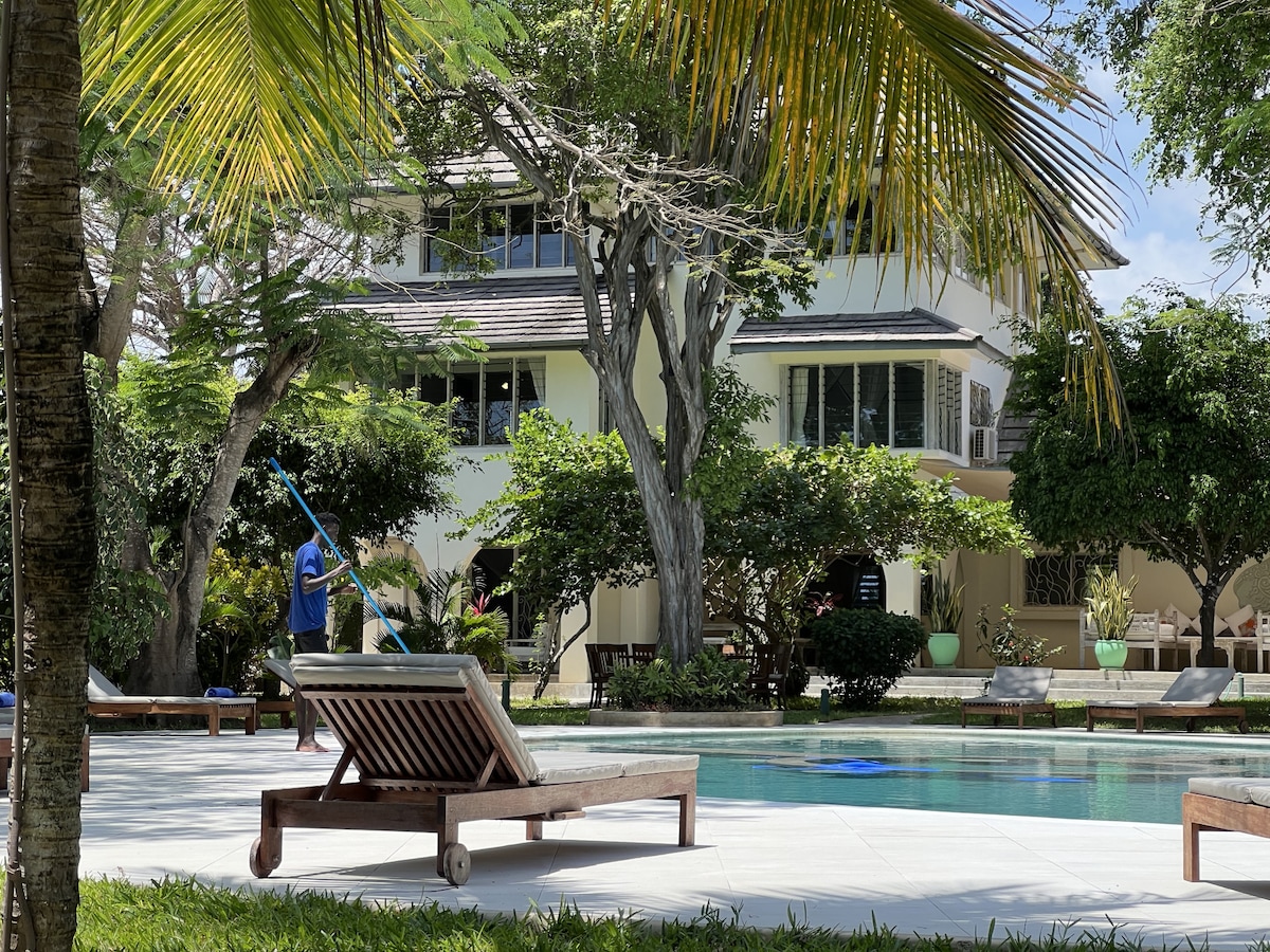 A serene pool area is set within a lush garden, featuring sunbeds arranged around the clear water. In the background, a multi-story villa is framed by vibrant trees, while a staff member is seen maintaining the pool's cleanliness.