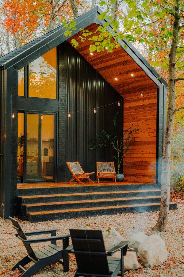 A modern cabin exterior is set against vibrant autumn foliage. The structure features large glass doors and warm wood accents, with a cozy deck showcasing two wooden chairs. A fire pit area with stones surrounds the base, adding to the inviting outdoor atmosphere.