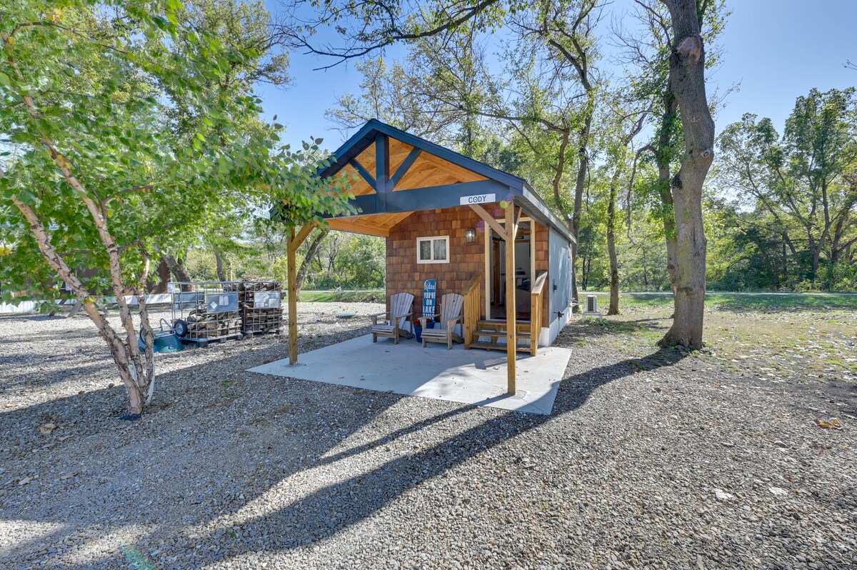 A single-story cabin is presented under a covered patio, featuring outdoor seating. The structure's exterior combines natural wood tones with a modern overhang. Surrounding greenery and gravel paths enhance the serene environment.