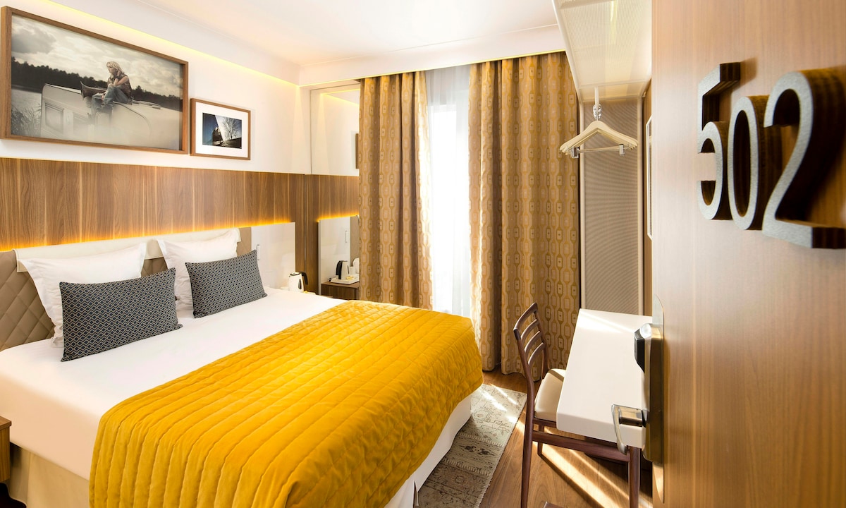A well-lit room features a spacious bed dressed in a vibrant yellow coverlet. Gray accent pillows add contrast. Natural light enters through a window, complemented by elegant drapes. A sleek desk and chair are present, while custom wall art and subtle lighting enhance the modern decor.