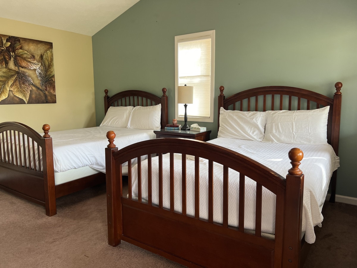 Two spacious beds are positioned on either side of a shared nightstand, with a lamp providing soft lighting. Green walls enhance the calming atmosphere, while a piece of artwork hangs above one of the beds, adding a touch of elegance.