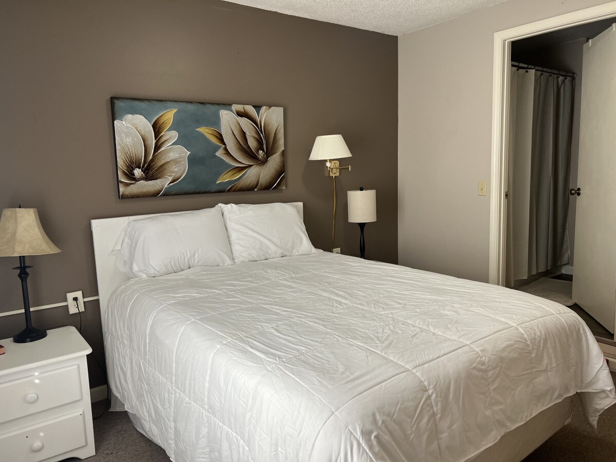 A bright bedroom features a spacious bed with white linens and a floral wall art piece above the headboard. Two light fixtures, one on a nightstand and another freestanding, provide illumination. A doorway on the right leads to a bathroom area, partially visible.