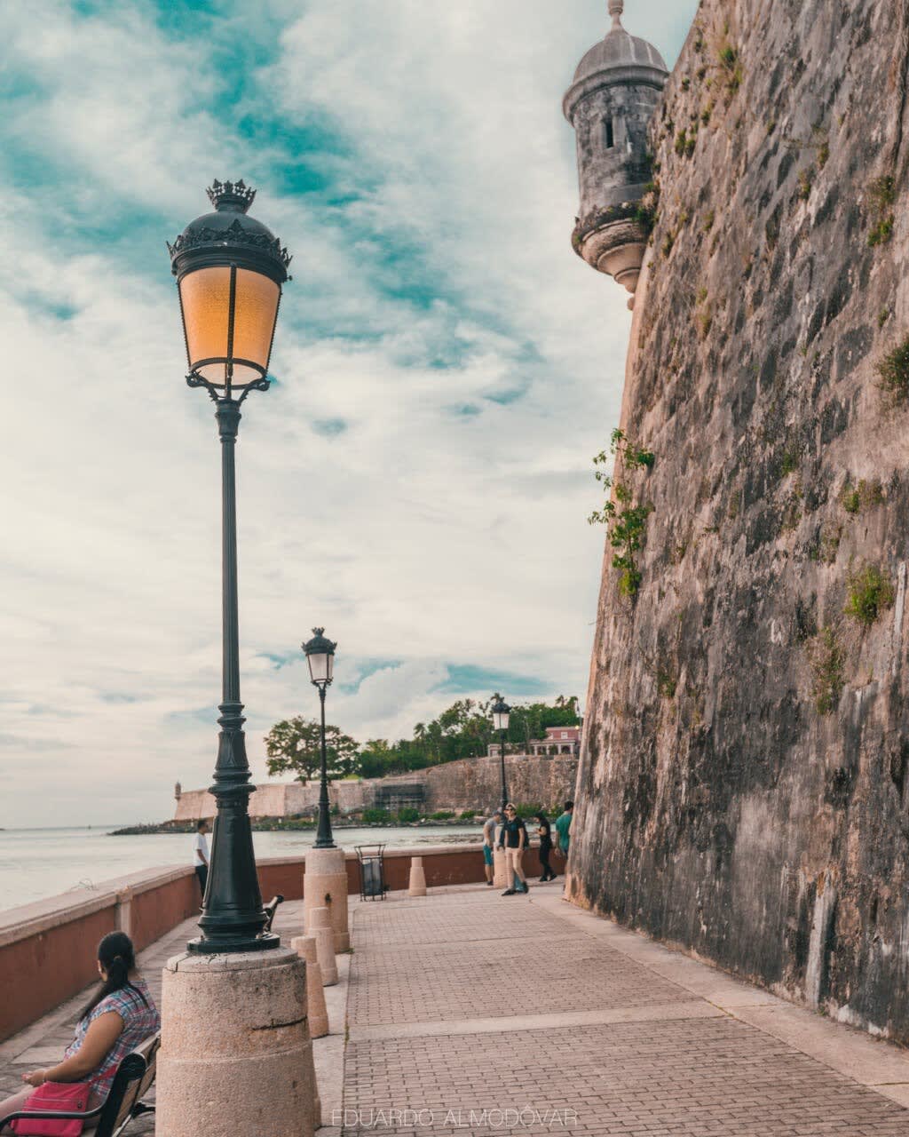 A charming waterfront walkway is lined with vintage street lamps, casting a warm glow against a backdrop of a historic stone wall. The serene sea is visible to one side, inviting peaceful strolls along this scenic path.
