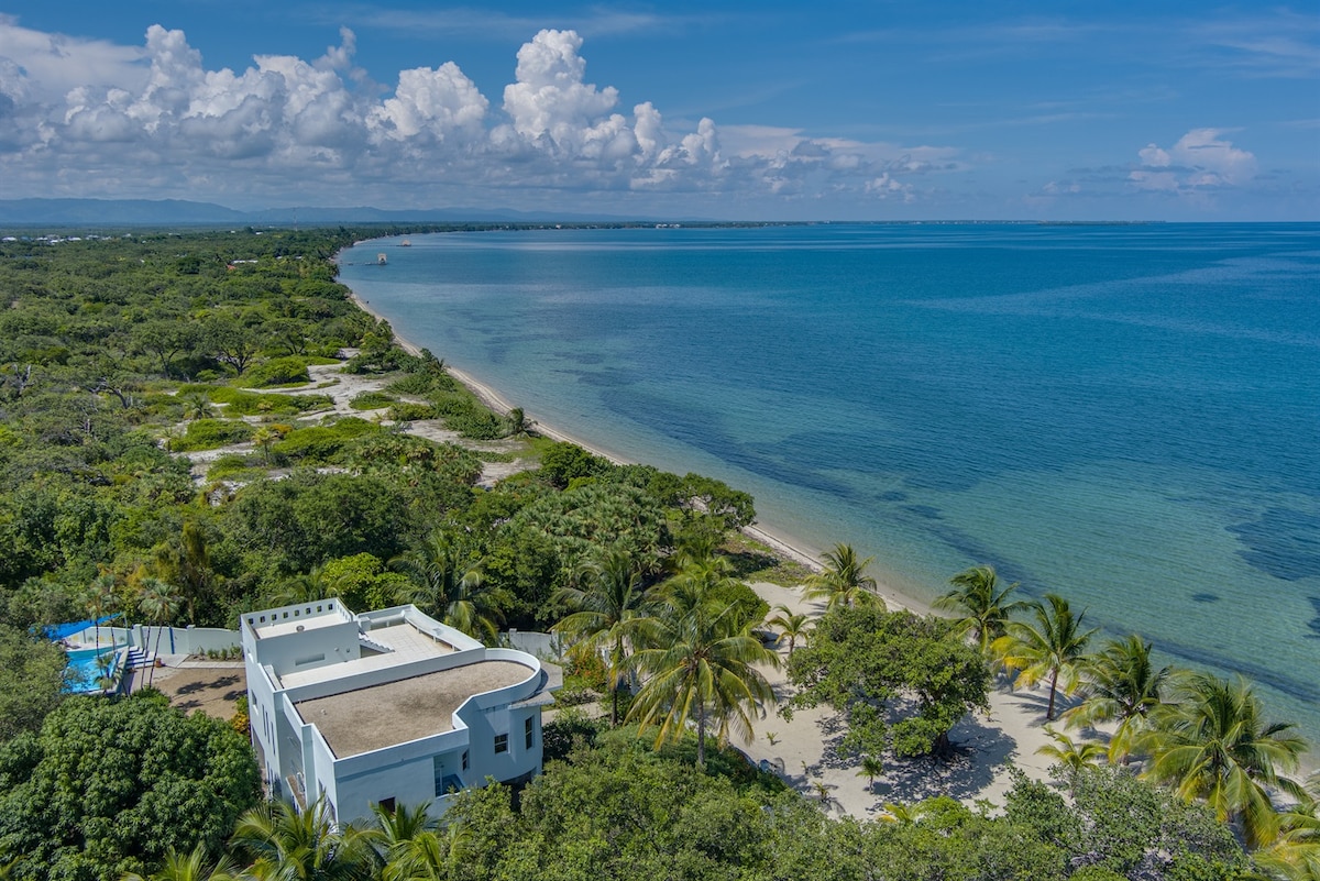 An aerial view captures the serene coastline of Placencia, Belize, featuring clear blue waters and sandy beaches framed by lush tropical foliage. The Casa Nettuno property is positioned gracefully along the shoreline, surrounded by vibrant greenery, offering a sense of seclusion and tranquility.