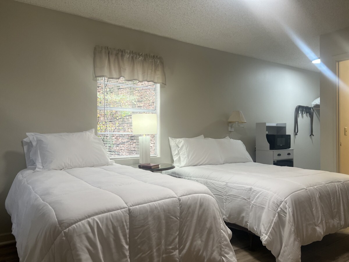 A well-lit bedroom is furnished with two spacious beds dressed in white linens. Natural light enters through a window, beside which a lamp and a small shelf are placed. A microwave is visible on a stand across the room.