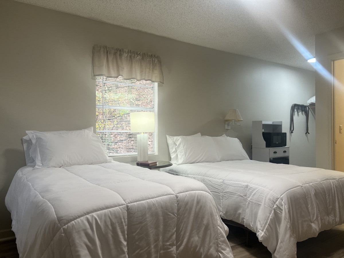 This sleeping area features two spacious beds, each dressed in white bedding. A window allows natural light, complemented by a bedside lamp. A microwave and small refrigerator are positioned nearby.