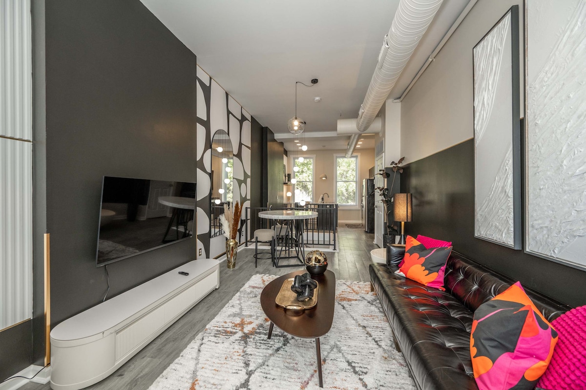 A modern living area showcases a plush leather sofa with colorful accent pillows. A sleek TV is mounted on the wall opposite a low-profile coffee table. Behind, a dining area is visible, highlighted by large windows that allow natural light to fill the space.