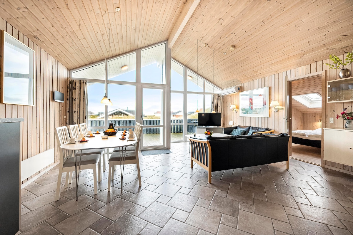 A spacious living area is defined by high wooden ceilings and large windows that fill the room with natural light. A dining table with white chairs is set for meals, and a comfortable sofa provides a gathering space. A TV is visible in the background.