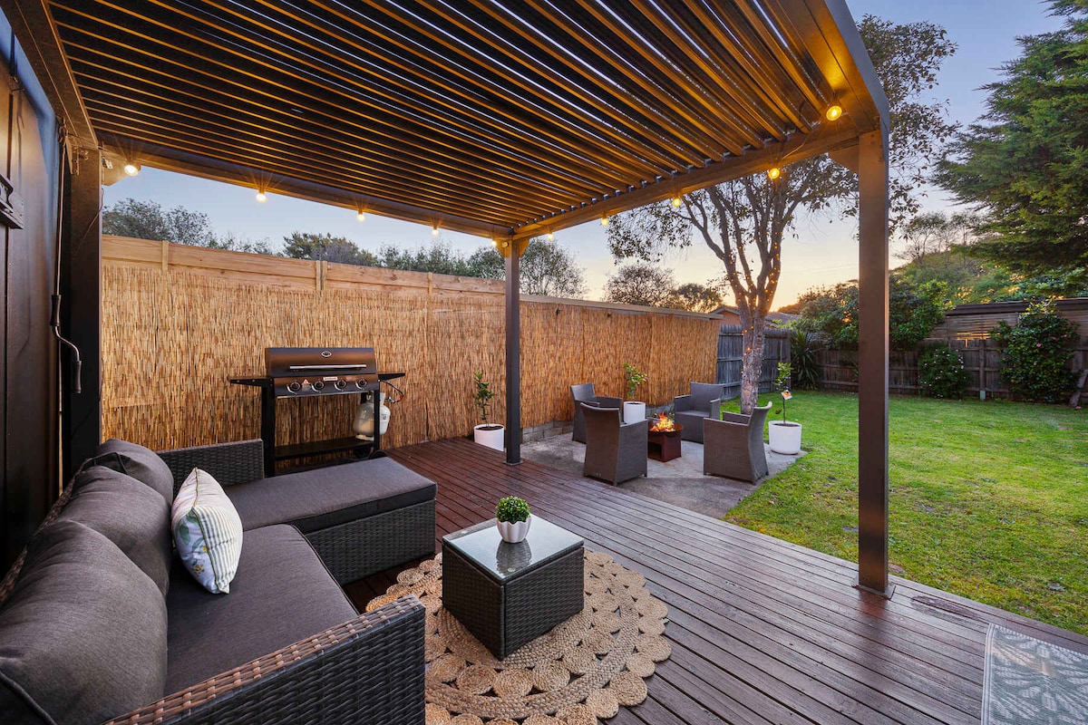 An outdoor entertaining area features a covered lounge setup with dark wicker furniture, a circular rug, and soft lighting. A BBQ grill is positioned nearby, complemented by a fire pit surrounded by white planters. The green lawn provides an inviting space for gatherings.