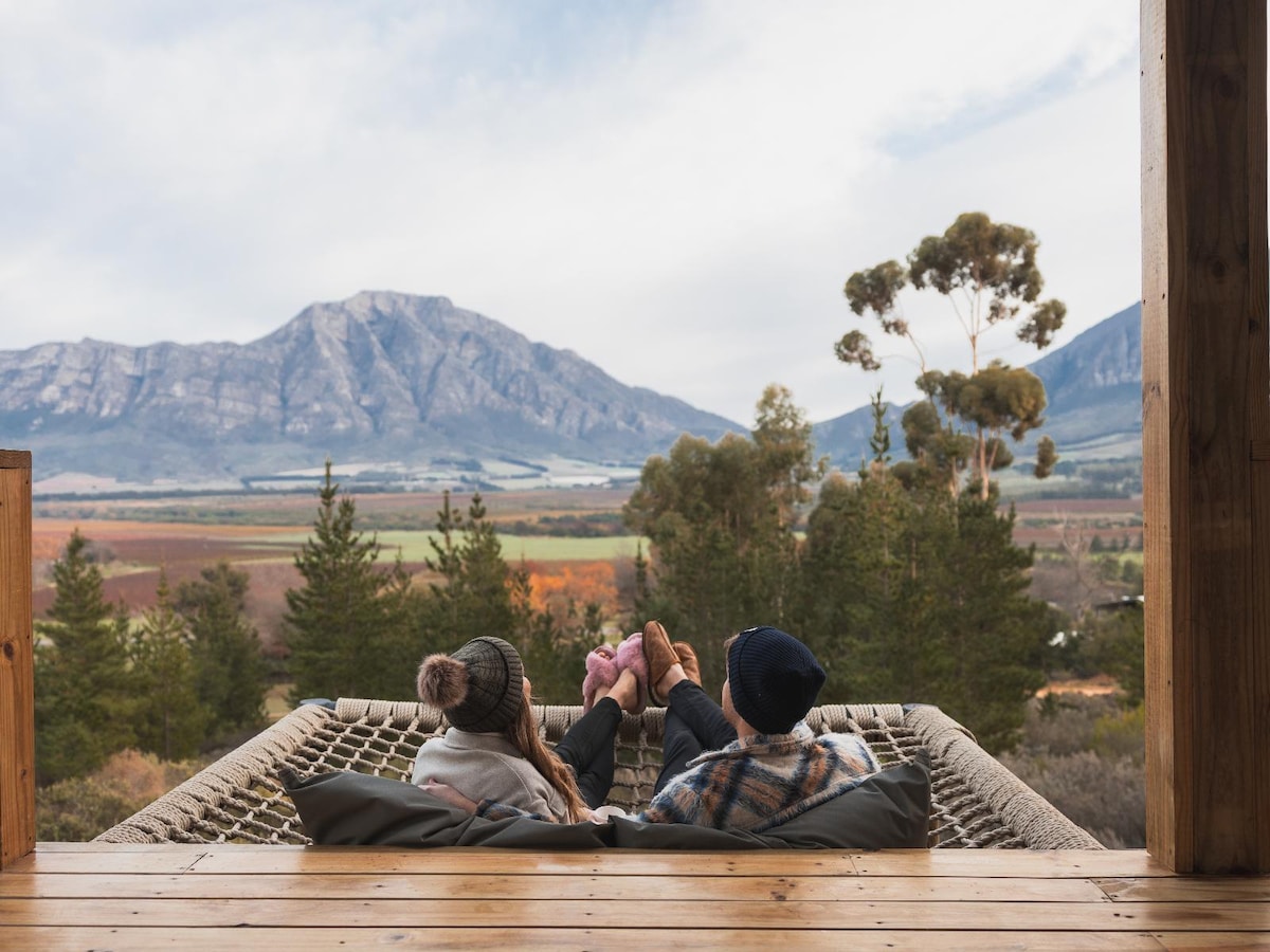 Two individuals are seated on a comfortable hammock, enjoying a scenic view of mountains and rolling hills. Soft earth tones are present in the surrounding nature, while tall trees frame the landscape, creating an inviting setting for relaxation.