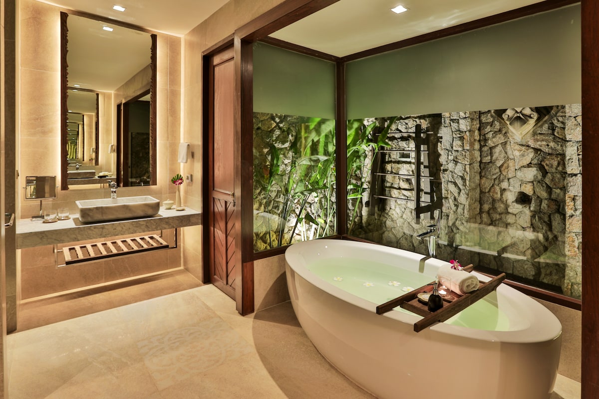 A modern bathroom features a freestanding tub elegantly positioned near large windows, providing views of a natural stone wall and lush greenery. A sleek vanity with a basin completes the space, enhancing the spa-like ambiance.