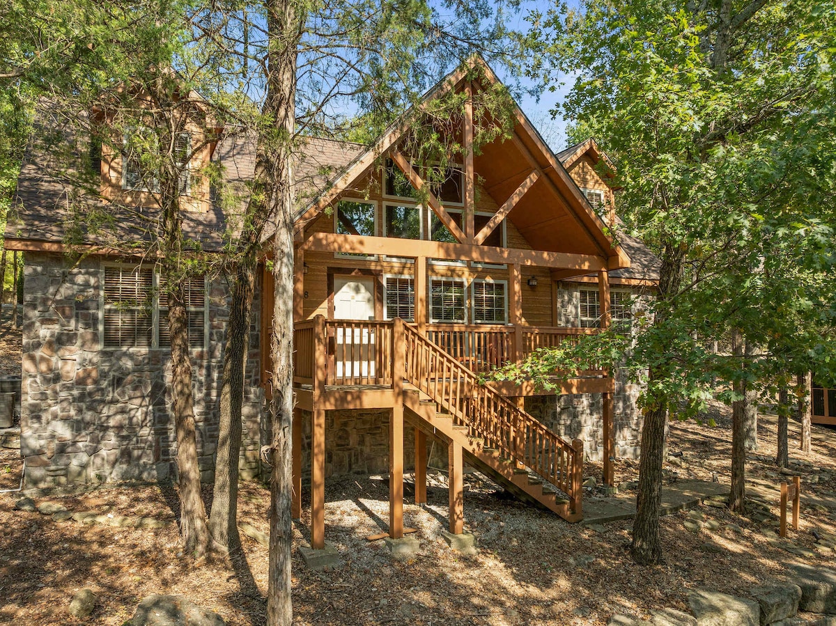 A rustic cabin exterior is surrounded by lush greenery, with tall trees providing shade. The wooden front deck features a staircase leading to the entrance and large windows allow for natural light. Stone and wood accents contribute to the warm, inviting look of the home.
