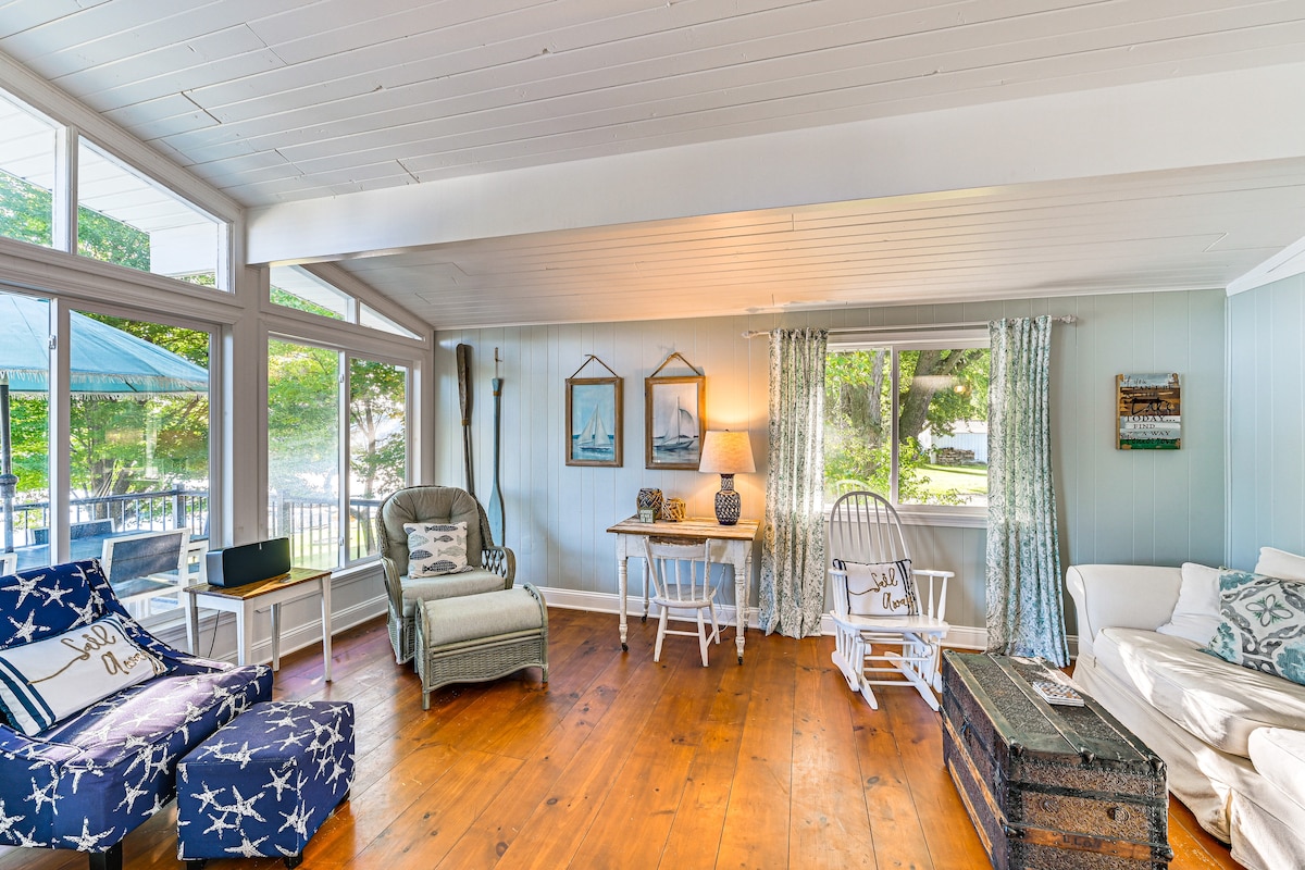 A bright living space showcases large windows allowing natural light to fill the room. Comfortable seating includes a rocking chair and patterned armchairs. A small table and decorative items complement the relaxed atmosphere. Wooden flooring adds warmth, while light-colored walls enhance the airy feel.