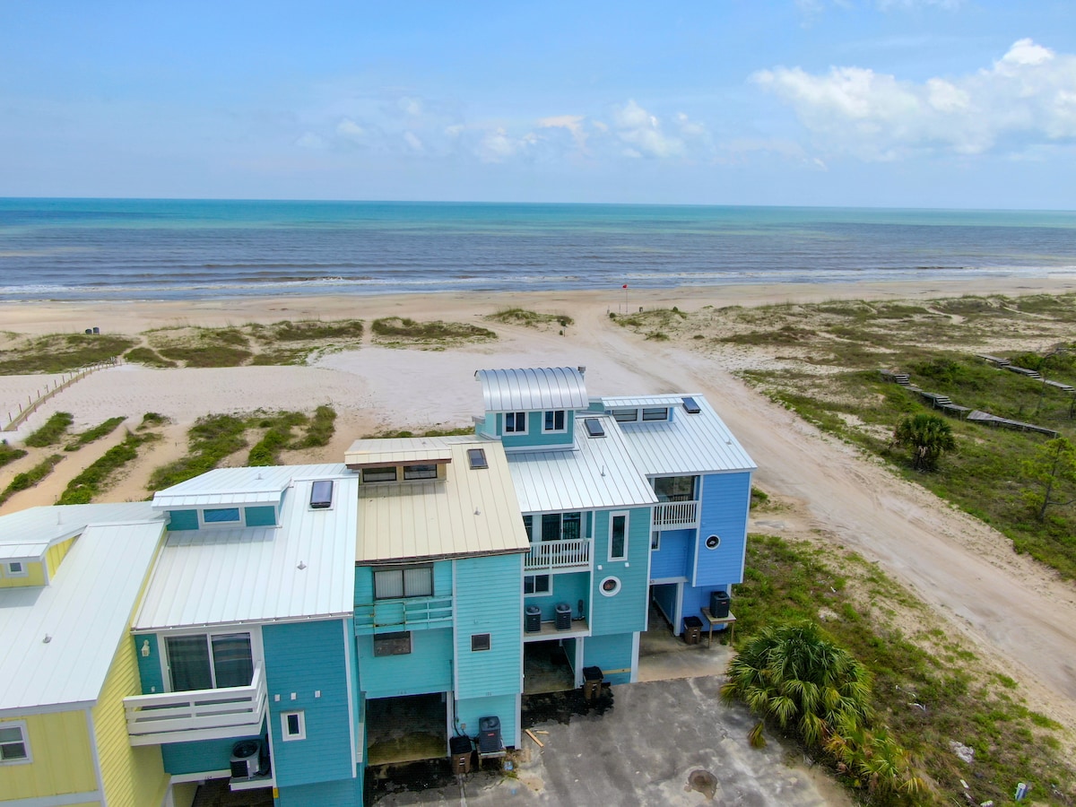 An aerial view of the Gulf Front Townhome showcases its bright colors against the sandy beach and turquoise waters of the Gulf of Mexico. A clear sky and gentle waves create a serene coastal atmosphere. The surrounding area features an expanse of sand and greenery.