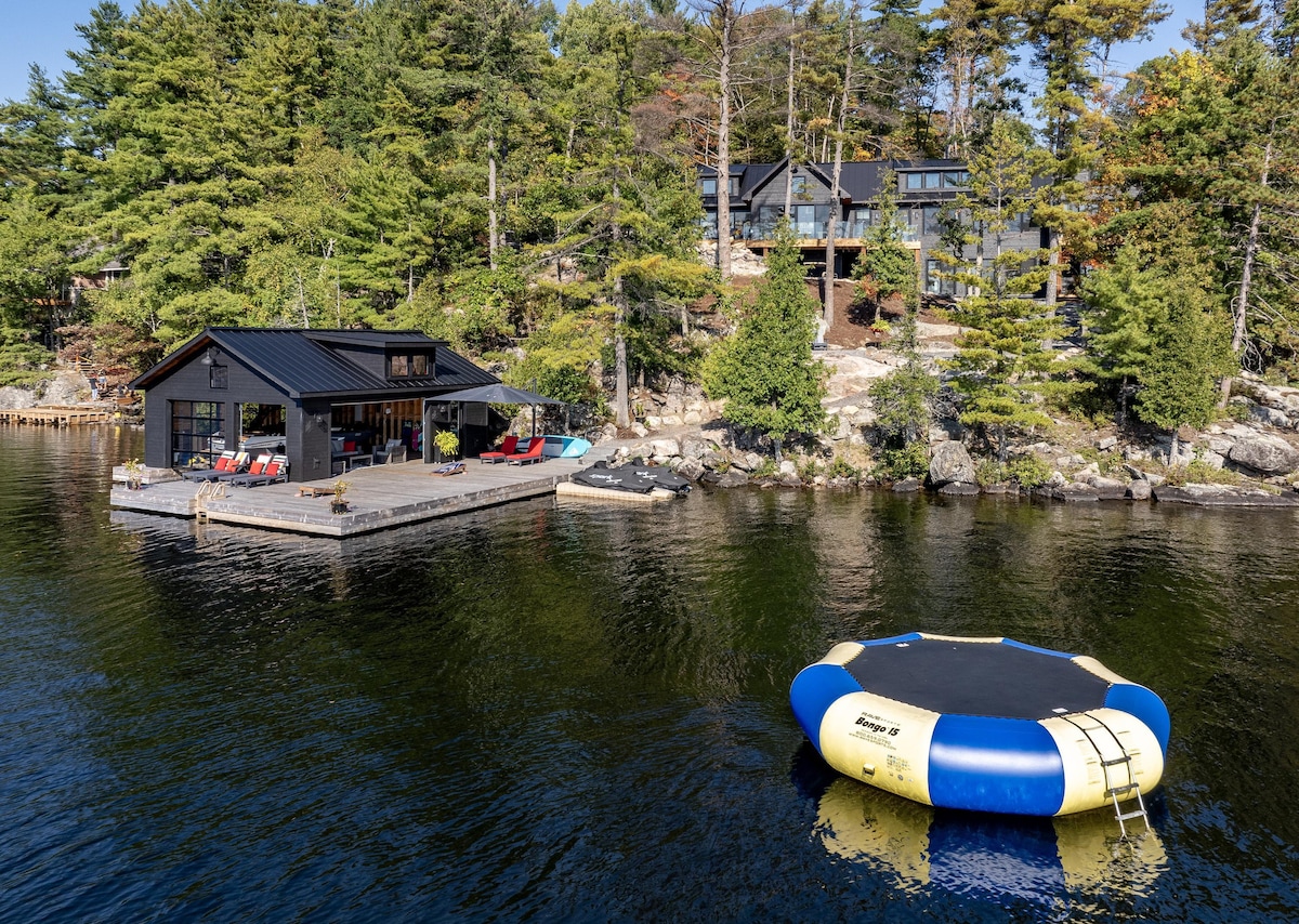A modern boathouse is positioned on the tranquil waters of Lake Rosseau, with a large water trampoline in the foreground. Towering trees surround the property, providing a natural backdrop, while a spacious deck showcases various seating options and direct access to the lake.