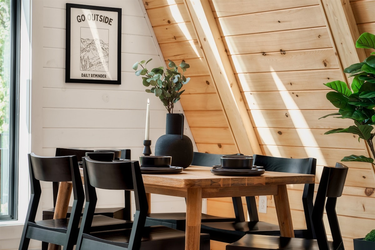 A modern dining area features a rectangular wooden table with black plates and sleek chairs arranged around it. Natural light filters in through a nearby window, illuminating the wood-paneled wall and a framed motivational print. A decorative plant and a candle serve as centerpieces.