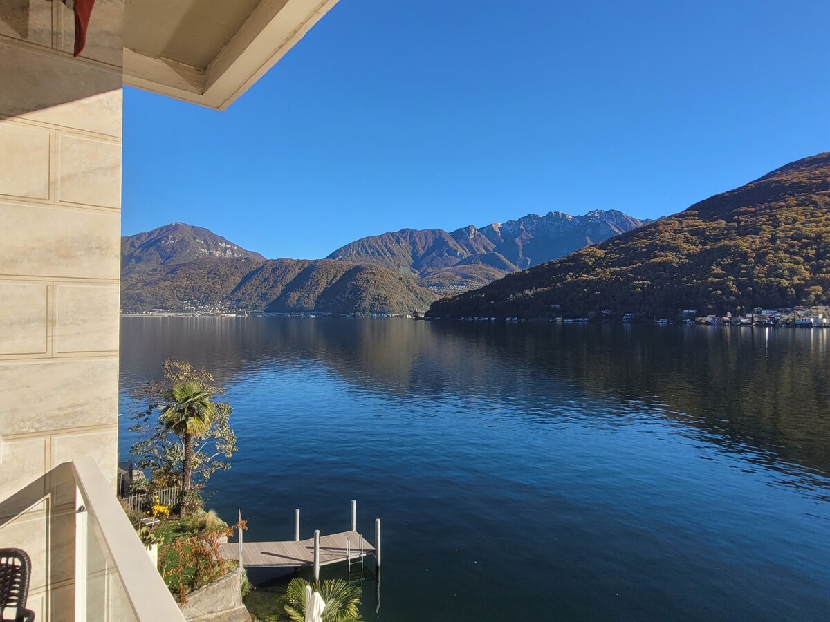 A serene view of Lake Lugano is visible from the balcony, showcasing calm waters reflecting the surrounding mountains. Clear blue skies enhance the peaceful atmosphere, while a small dock is positioned along the shoreline, contributing to the picturesque lakeside setting.