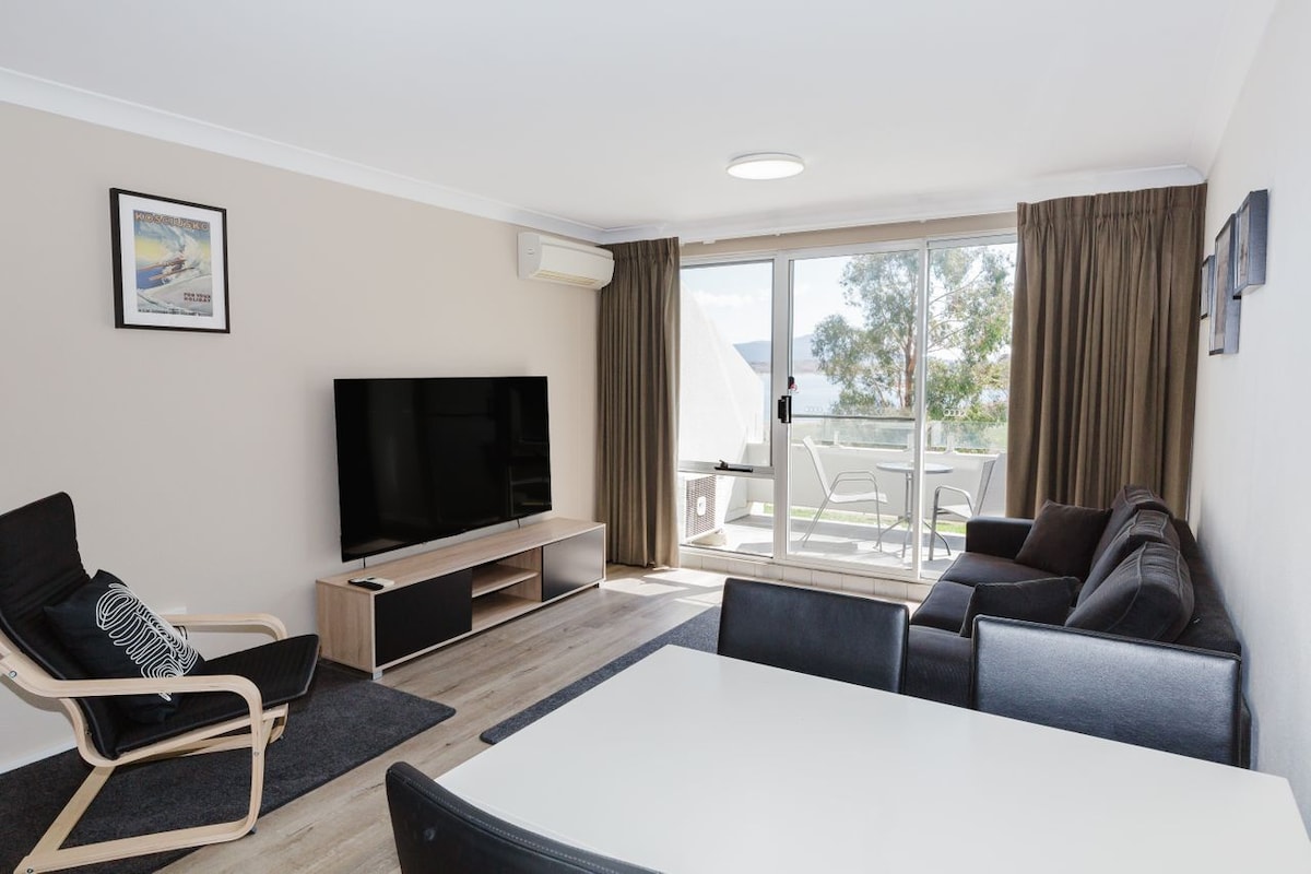 A contemporary living area features a large flat-screen television mounted on a light wooden console. A dark sofa and a black dining table accompany a light-toned armchair with a minimalist design. Floor-to-ceiling glass doors provide access to a balcony with seating, offering scenic views.