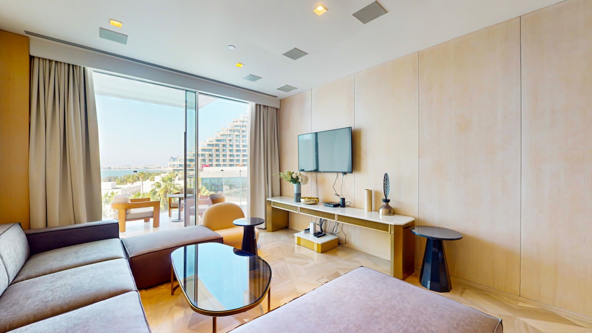 An open-plan living area features a comfortable sofa and an elegant lounge chair facing a Smart TV. A glass coffee table is positioned on a marble floor, and floor-to-ceiling windows provide ample natural light and views of the outdoor balcony.