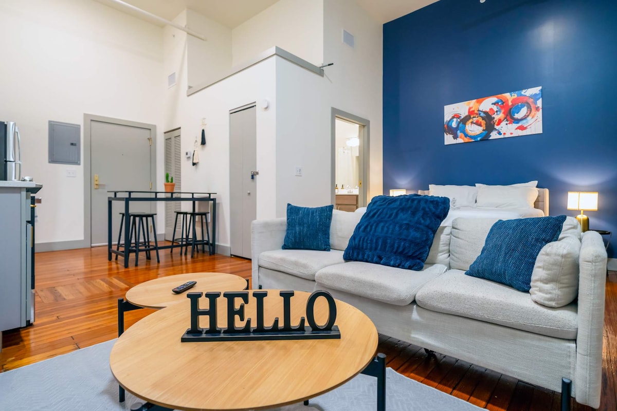 The spacious studio features a light-colored sofa adorned with navy pillows, positioned adjacent to two wooden coffee tables. A dark blue accent wall complements the area, while a dining table with high stools is visible in the background, leading to a bright kitchen area.