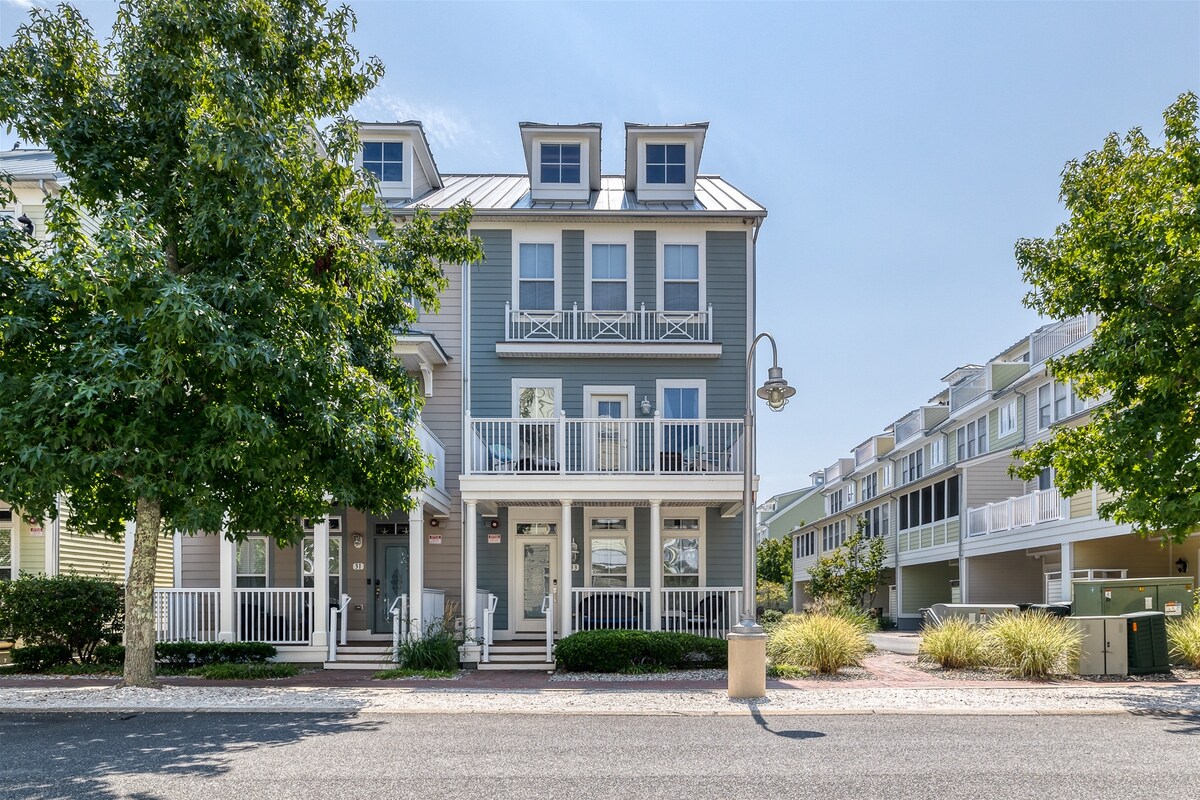 A charming townhouse exterior is presented, featuring a light blue facade with multiple levels. Two private balconies are visible on the front, framed by greenery from surrounding trees. The entrance is accessible from a small porch with steps, contributing to the inviting atmosphere.