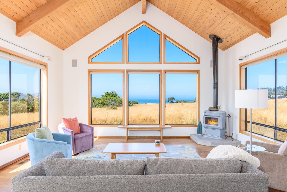 The spacious living area features a high, wood-paneled ceiling and large windows that frame views of the coastline and surrounding landscape. A comfortable sofa and armchairs are arranged around a central coffee table, and a modern wood stove adds warmth to the space.
