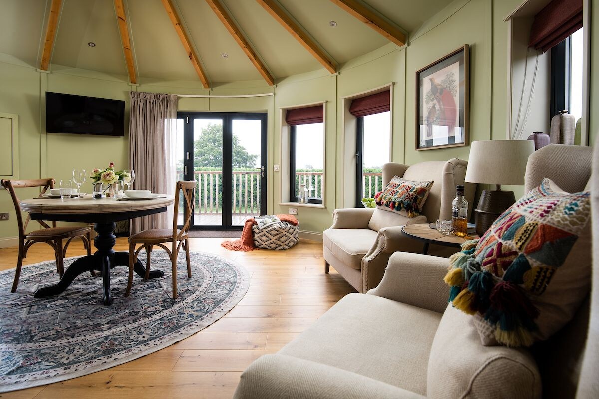 An open-plan living area is highlighted by natural light from the full-height windows and doors. A round dining table for two is set near the center, surrounded by cozy seating options. Decorative cushions add a touch of color to the neutral-toned armchairs.